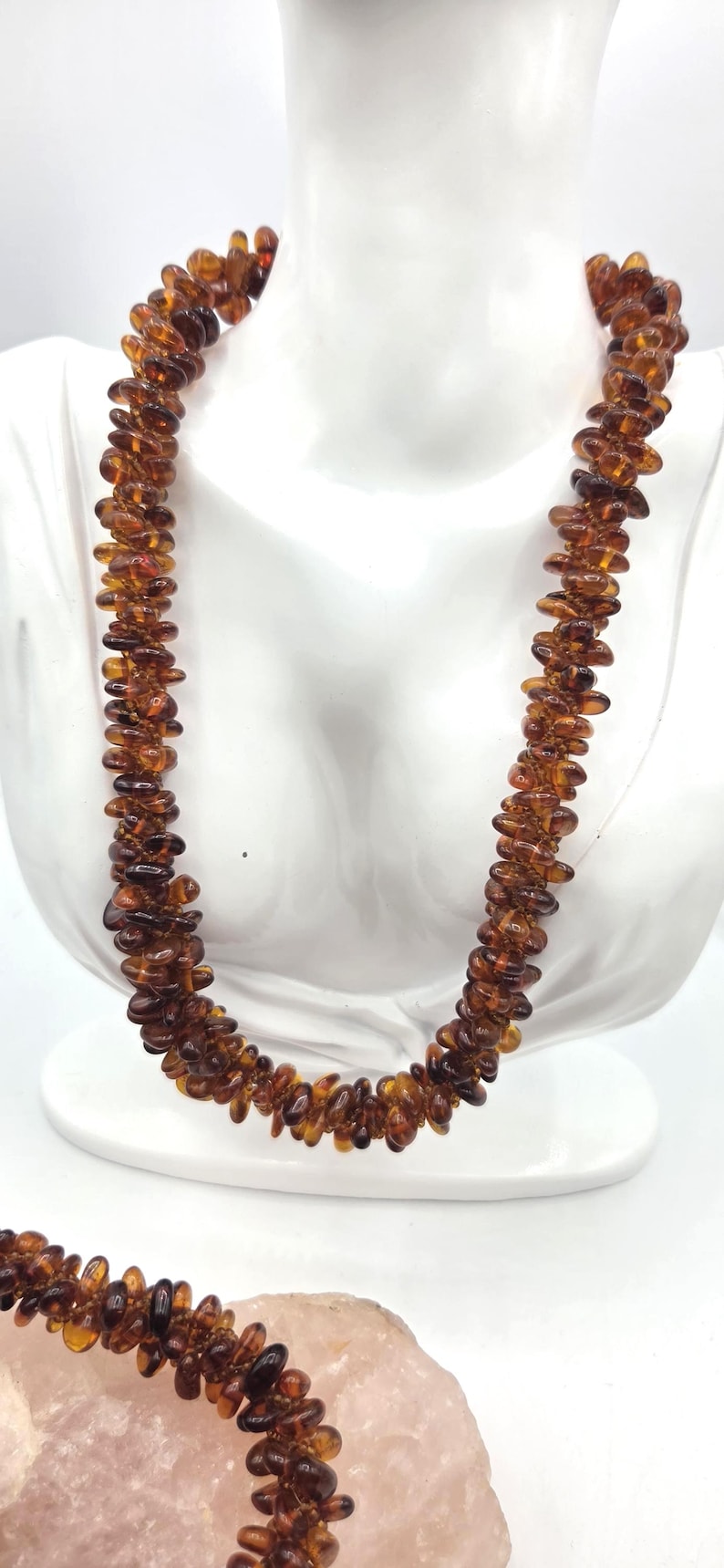 May include: A long necklace composed of numerous small, polished, translucent amber beads in varying shades of deep cognac and honey brown. The beads are strung together in a dense, multi-layered arrangement, creating a textured, organic appearance.