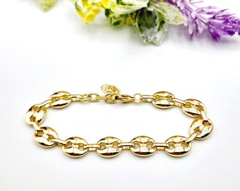 May include: A gold-colored bracelet with a series of oval-shaped links, connected by smaller circular links. The bracelet is displayed on a white surface, with a blurred background of flowers in yellow, green, and purple.