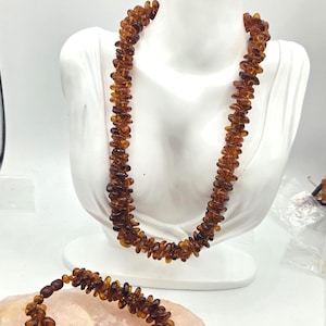 May include: A necklace and a bracelet set composed of numerous small, polished, amber-colored resin beads. The necklace is draped over a white display bust, while the matching bracelet is positioned separately in the foreground.