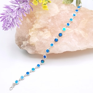 May include: A silver bracelet featuring a series of round, blue opal stones. The bracelet is displayed on a light pink stone with purple and yellow flowers in the background. The opal stones vary in shades of blue.