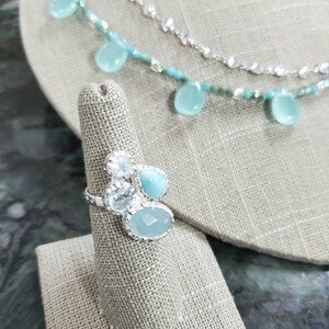Puede incluir: Un anillo de plata con un grupo de piedras preciosas azules y transparentes. El anillo se muestra en un soporte de anillo de tela.