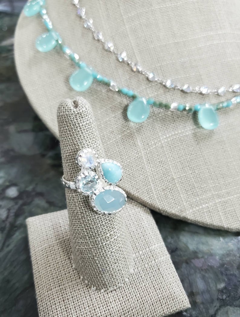 Puede incluir: Un anillo de plata con un grupo de piedras preciosas azules y transparentes. El anillo se muestra en un soporte de anillo de tela.