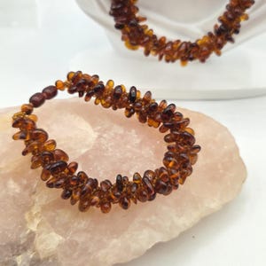 May include: A bracelet and a necklace made of small, polished, translucent amber beads in shades of deep cognac and honey brown. The beads are irregularly shaped and strung together to create a textured, multi-layered appearance.