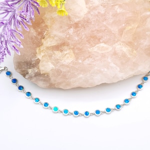 May include: A silver bracelet featuring a series of round, light blue opal stones. The stones are set in a delicate silver chain. The bracelet is displayed against a light pink stone and floral accents.