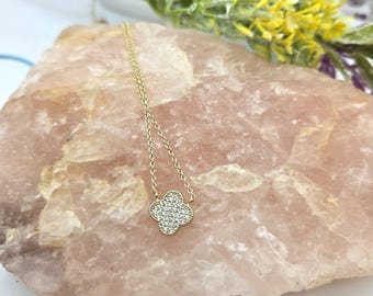 May include: A gold-colored necklace with a four-leaf clover pendant. The pendant is encrusted with small, clear gemstones. The necklace is displayed on a light pink, textured surface, with greenery and yellow flowers in the background.