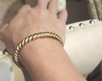 May include: A polished gold-tone bangle bracelet featuring a continuous twisted rope design. The circular metal band has a smooth, reflective surface with a repeating spiral pattern that wraps around the entire circumference of the jewelry piece.