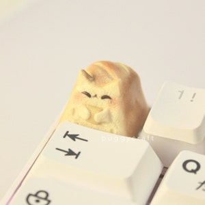 May include: A custom keycap featuring a cat design. The keycap is light brown with a smiling cat face and is placed on a white keyboard. The keycap has a 3D appearance and is a unique accessory for mechanical keyboards.