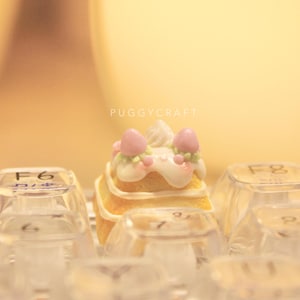 May include: A miniature cake keycap with pink and white icing, topped with pink and green decorations. The keycap is sitting on a keyboard with other keycaps.