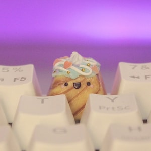 May include: A small, cute, and colorful cake-shaped keycap with a happy face and a swirl design. The keycap is sitting on a white keyboard with keys labeled "F5", "T", "Y", and "PrtSc".