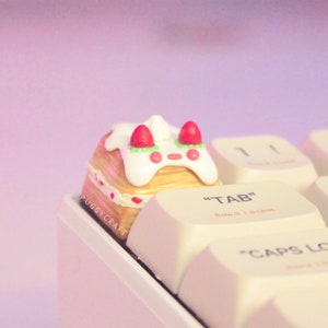 May include: A decorative keycap with a dessert design, featuring white icing and red strawberry details. The keycap is part of a keyboard, with the "TAB" key in view. The keycap has the text "PUGGYCRAFT" on the side.