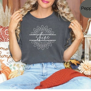 May include: A dark gray t-shirt with a white sunflower graphic and the word "Shine" written across the middle. The model is wearing blue jeans and has long blonde hair.