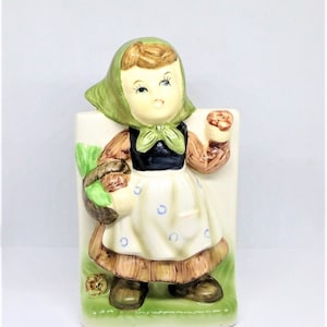 May include: A ceramic figurine of a girl wearing a green hooded dress and holding a brown object. She is standing on a green base with brown accents.