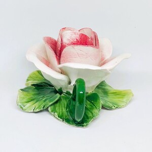 May include: A ceramic rose-shaped teacup with a green handle and green leaves. The rose petals are white with red edges. The teacup is on a white background.