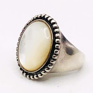 May include: A silver ring featuring an oval, off-white gemstone. The gemstone is set within a beaded border. The ring has a simple, polished band. The jewelry is displayed against a white background.