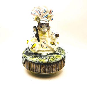 Puede incluir: Figura de cerámica de un ratón vestido con un vestido blanco y un gran sombrero con plumas, sosteniendo una lupa. El ratón está sentado sobre una base redonda decorativa con un patrón de vetas de madera.