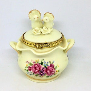 May include: A white porcelain trinket box with a floral design and a lid featuring two white doves. The lid has a gold trim and the box has two handles.