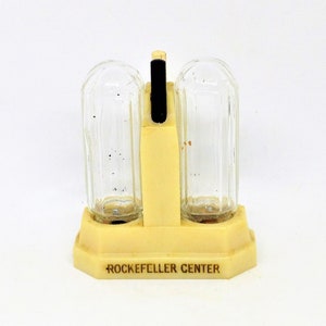 May include: A vintage salt and pepper shaker set with a cream-colored base and clear glass shakers. The base has the text "Rockefeller Center" printed on it.