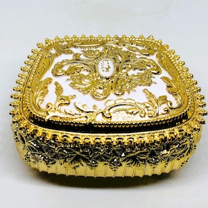 May include: A gold-colored metal trinket box with an ornate design. The box has a white interior with gold accents and a small, clear jewel in the center.