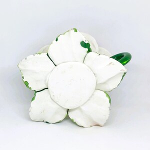 May include: A white, flower-shaped decorative object with a green glass handle. The flower has five large petals and a central disc, all painted white with green accents on the edges. The object is likely ceramic or plaster.