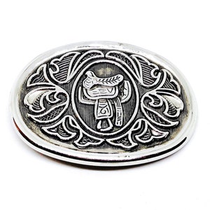 Puede incluir: Hebilla de cinturón plateada con un diseño grabado de una silla de montar. La hebilla tiene forma ovalada y presenta un patrón floral detallado alrededor de la silla de montar.