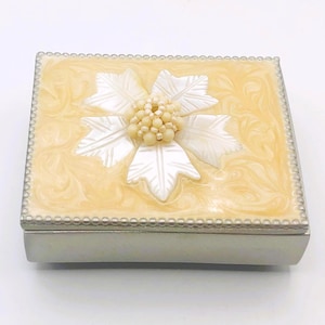 Peut inclure: Une boîte rectangulaire en métal avec une base argentée et un couvercle doté d'une finition en émail marbré crème. Le centre du couvercle est orné d'un motif floral blanc sculpté et d'un groupe de petites perles rondes couleur crème.
