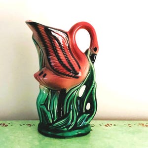 Puede incluir: Una jarra de cerámica con forma de flamenco y acabado brillante. El cuerpo de la jarra es de un vibrante color rosa coral con detalles de alas en negro, mientras que la base presenta follaje verde esculpido. El asa forma el cuello.