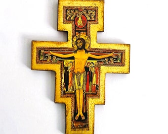 May include: A wooden wall cross featuring a gold-painted frame and a central painted icon of the crucified figure. The artwork includes smaller figures flanking the central image and a circular motif at the top, all rendered in a flat, stylized manner.