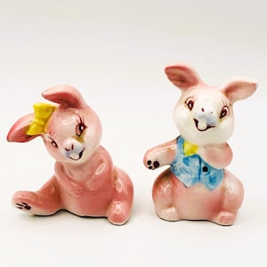 May include: A pair of pink ceramic bunny salt and pepper shakers. The female bunny has a yellow bow and the male bunny has a blue vest.