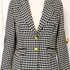 May include: A black and white houndstooth blazer with gold buttons. The jacket has a classic notched collar, two front pockets, and a tailored fit. The pattern is a repeating design of small, irregular shapes.