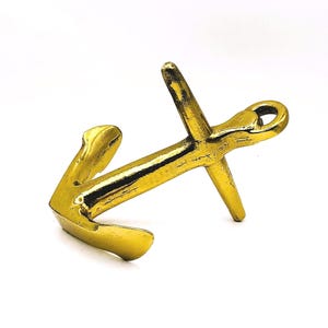 Puede incluir: Un ancla dorada con un diseño clásico, con una aleta curva y un anillo en la parte superior. El ancla está hecha de un material metálico brillante y está colocada sobre un fondo blanco. Este artículo de temática náutica es ideal para la decoración del hogar.