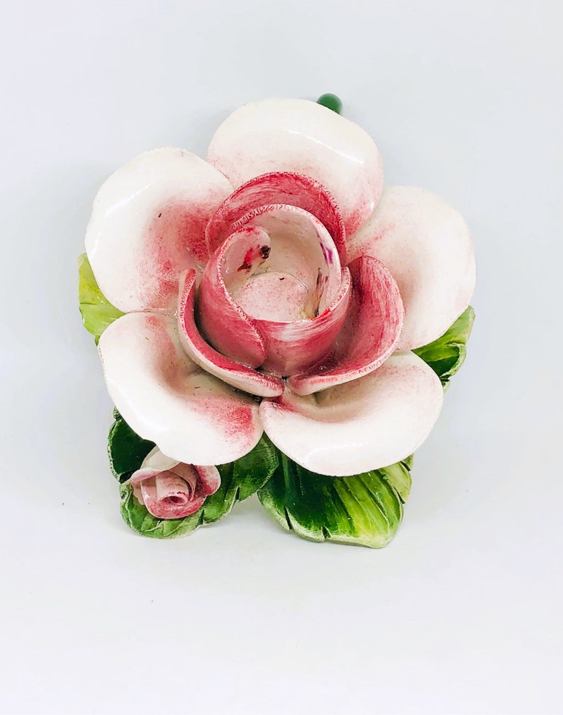 May include: A ceramic rose with pink and white petals and green leaves. The flower has a small bud and a green stem. The petals have a glossy finish. The rose is a decorative item.