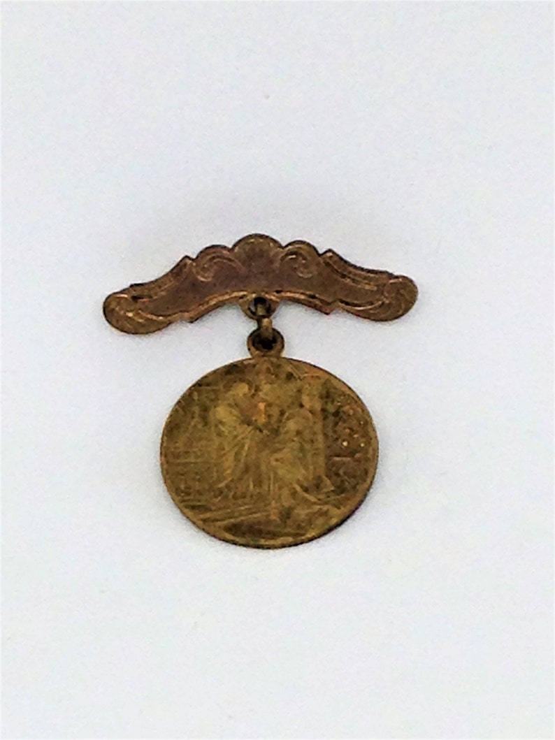 May include: A gold-colored metal pin with a round, embossed design and a curved, decorative top.