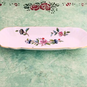May include: A white, rectangular porcelain dish with a scalloped edge and gold trim. The dish is decorated with colorful floral patterns in pink, blue, and green. The dish is approximately 10 inches long.