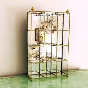 Puede incluir: Una vitrina rectangular dorada con múltiples estantes de vidrio. El mueble tiene una estructura de rejilla con respaldo de espejo y está rematado con pináculos decorativos. El mueble está colocado sobre una superficie verde.