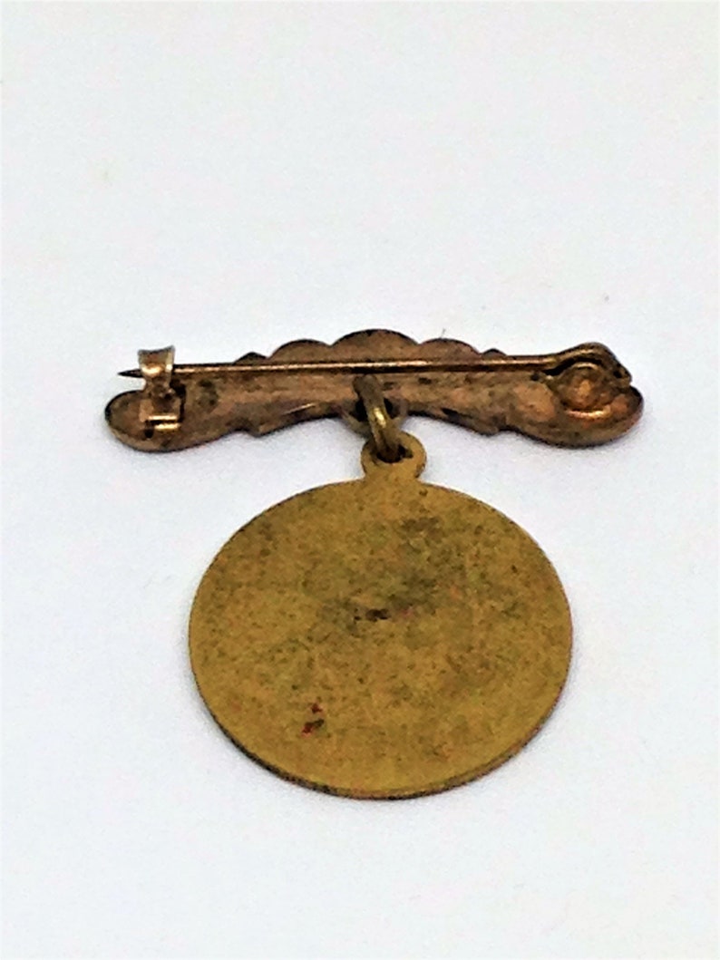 May include: A gold-toned pin with a round, blank, metal charm hanging from it. The pin is in the shape of a curved line with a safety clasp.