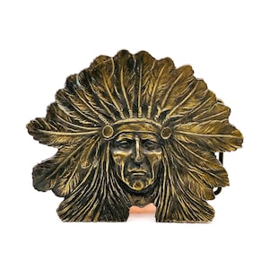 May include: A bronze-colored belt buckle featuring a detailed depiction of a Native American chief wearing a feathered headdress. The buckle has a rectangular shape with a central portrait and intricate feather detailing.