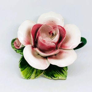 May include: A ceramic rose sculpture with white petals edged in pink and a deep pink center. The sculpture includes green leaves and a small bud. The flower is a decorative item.