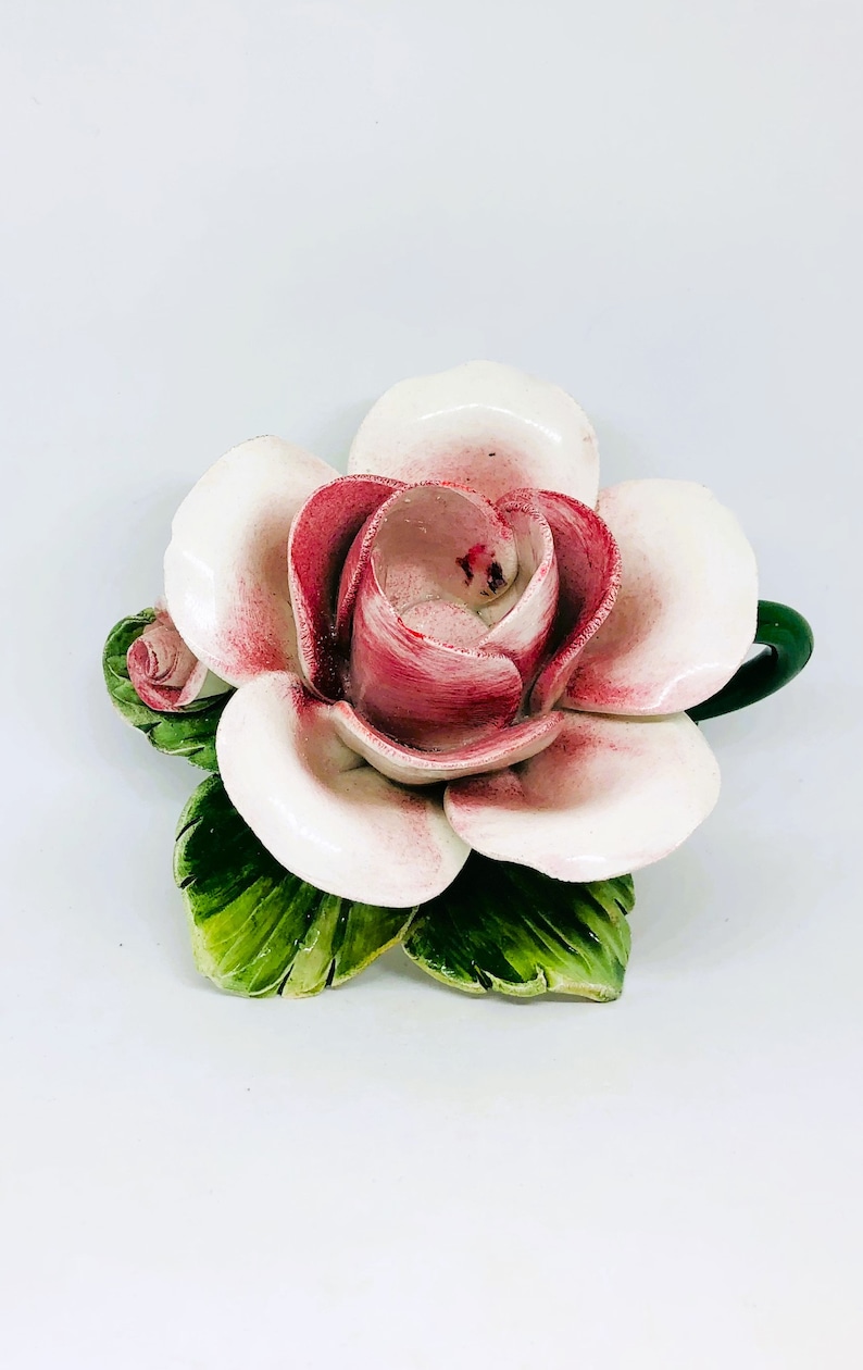 May include: A ceramic rose sculpture with white petals edged in pink and a deep pink center. The sculpture includes green leaves and a small bud. The flower is a decorative item.