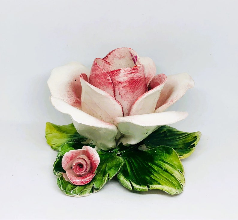 May include: A ceramic rose sculpture with a large, open bloom in shades of pink and white. The petals have a delicate, textured appearance. The sculpture includes green leaves and a smaller rose bud.