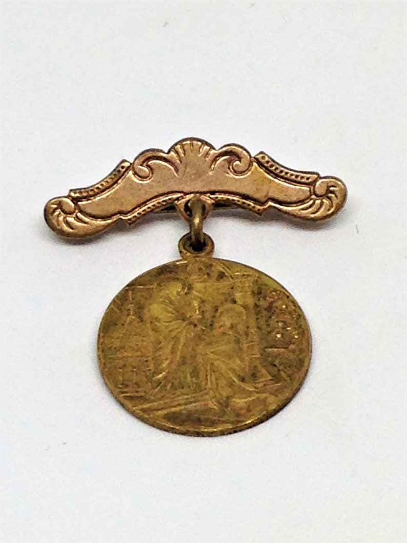 May include: A gold-toned pin with a decorative, ornate, curved top and a round, detailed medallion hanging from it.