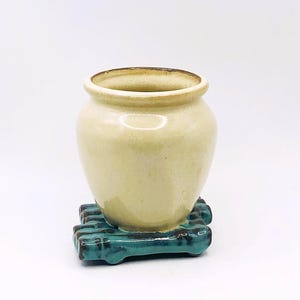 May include: A small, cream-coloured ceramic vase with a rounded body and a slightly flared rim. The vase sits on a square, teal-coloured ceramic base with a textured, rectangular pattern. The rim of the vase has a darker, brownish hue.
