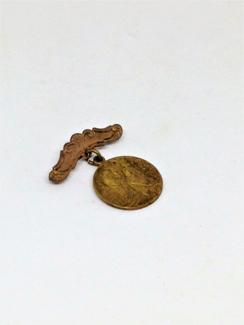 May include: A gold-colored metal pin with a round medallion featuring a detailed design. The pin has a decorative, curved bar.