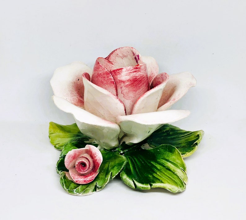 May include: A ceramic rose sculpture with a large, open bloom in shades of pink and white, accented with green leaves. A smaller rose bud is also present. The sculpture is set against a white background.