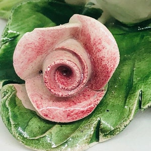 May include: A ceramic rose with pink petals and green leaves. The rose is the focal point, with detailed veining on the leaves. The piece appears to be a decorative item, possibly for a home decor.