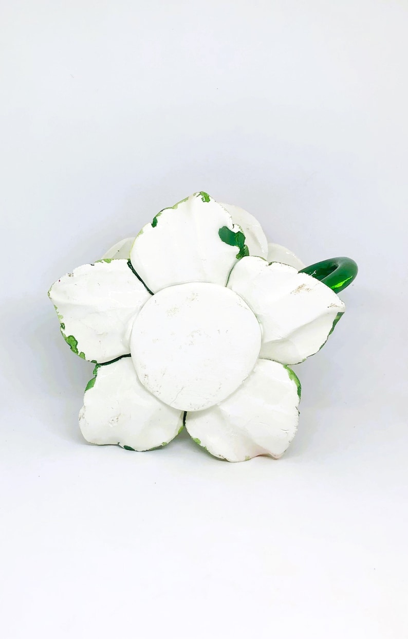 May include: A white, flower-shaped decorative object with a green glass handle. The flower has five large petals and a central disc, all painted white with green accents on the edges. The object is likely ceramic or plaster.