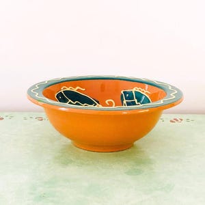 May include: Orange ceramic bowl with a teal and cream rim. The interior features teal fish designs. The bowl is round and sits on a light green surface. A decorative piece for the kitchen or dining room.