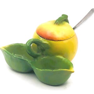 May include: A ceramic apple-shaped sugar bowl with a lid and a small spoon, accompanied by two leaf-shaped dishes. The sugar bowl is yellow with red accents, and the dishes are green. A charming kitchenware set.