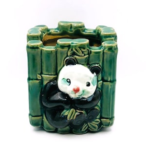 Könnte beinhalten: Keramik-Blumentopf mit Panda-Bär-Design. Der Topf ist grün mit einem Bambus-Außenbereich und einem Panda-Bären in Schwarz und Weiß mit roter Nase. Der Topf ist quadratisch.