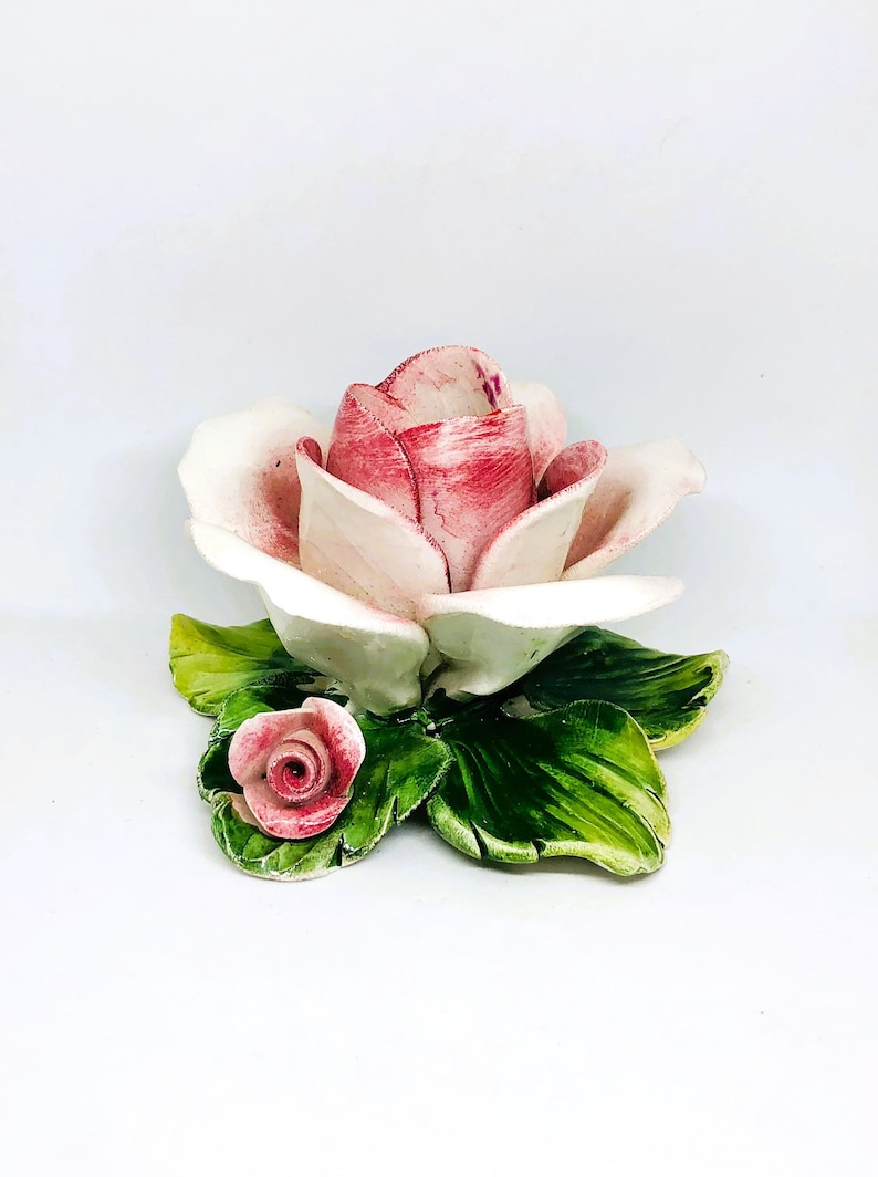 May include: A ceramic rose sculpture with a large, open bloom in shades of pink and white, accented by green leaves. A smaller rose bud is also present. The sculpture is set against a white background.