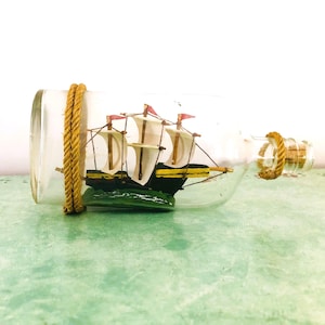 May include: A model sailing ship displayed inside a clear glass bottle. The ship features white sails, red flags, and a black hull with yellow detailing. The bottle is sealed with a cork and wrapped with gold rope.